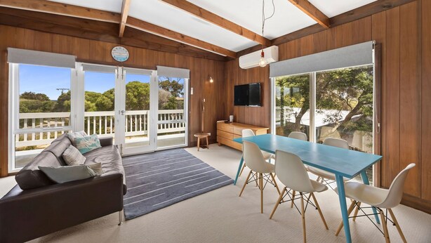 A spacious room with a wooden ceiling, a dining area with a blue table and white chairs, a sofa with cushions, a television mounted on the wall, and large sliding glass doors leading to an outdoor deck. - The Beach House on Sixth (Anglesea)