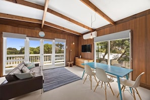 Ein geräumiger Raum mit Holzbalkendecke, einer Essecke mit blauem Tisch und weißen Stühlen, einer Couch mit Kissen, einem an der Wand befestigten Fernseher und großen Schiebeglasüren zu einer Außenterrasse.