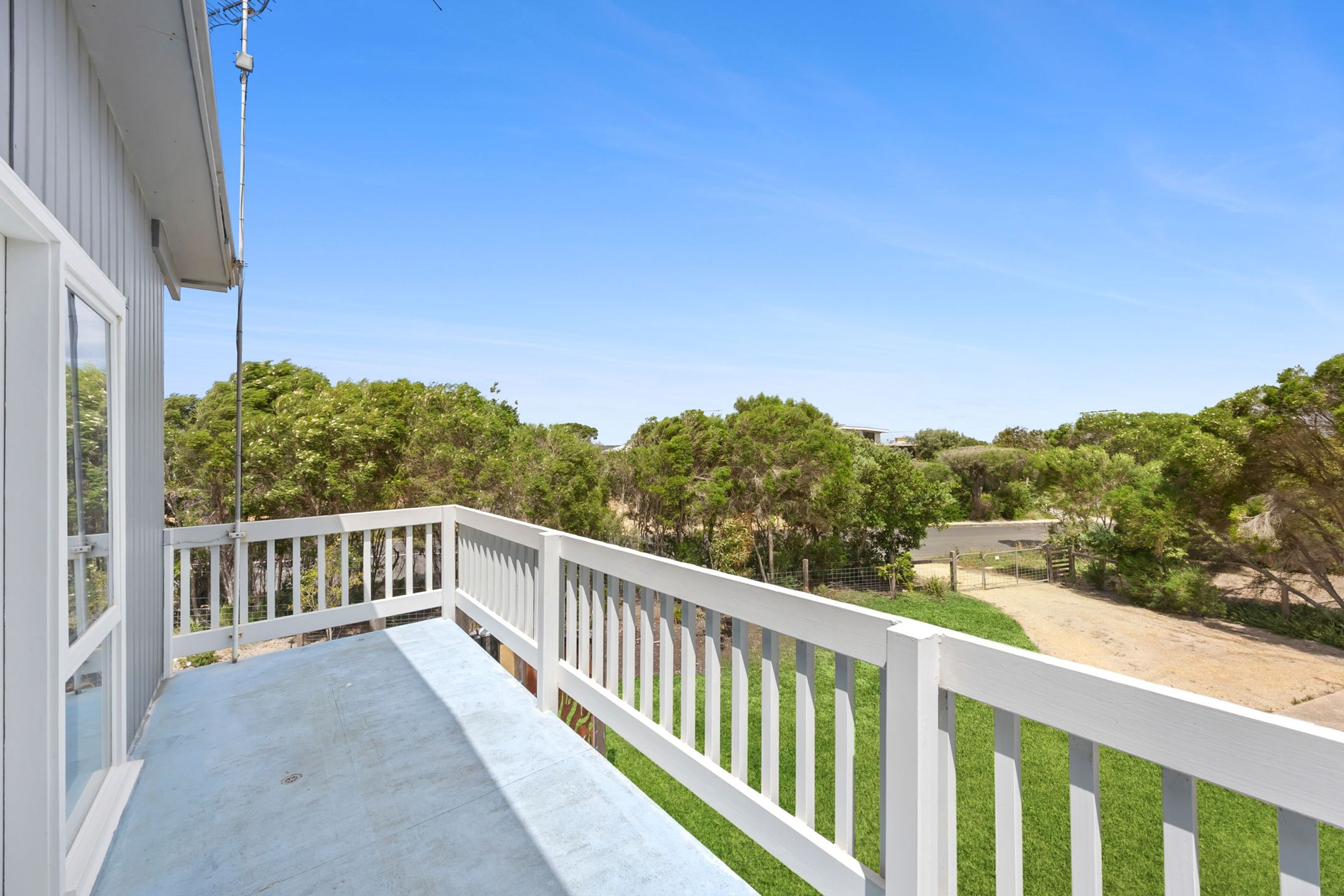A balcony with a white railing overlooking a grassy area with trees and a clear blue sky.