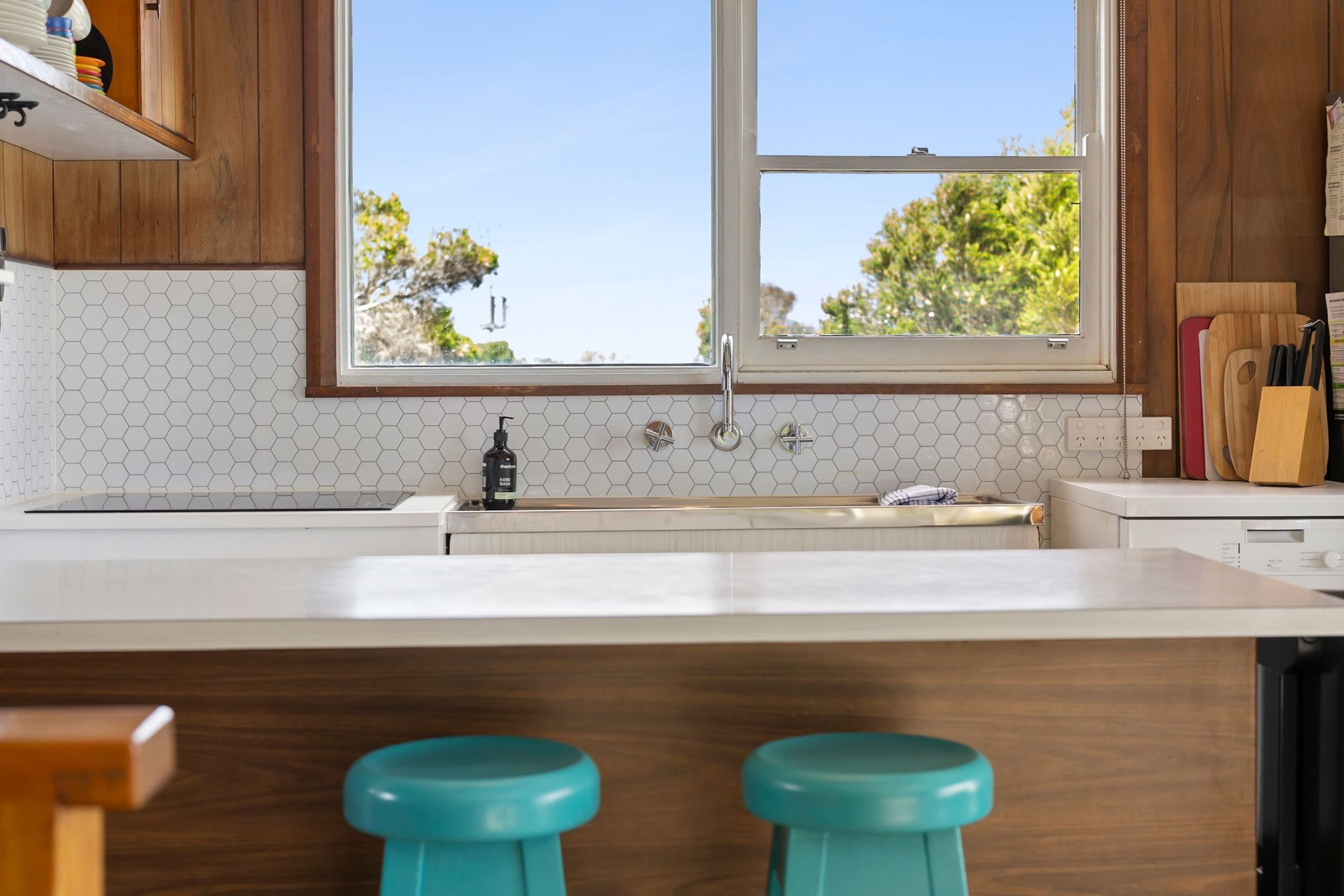 A kitchen with a large window, wooden cabinets, a white countertop, and two teal stools.