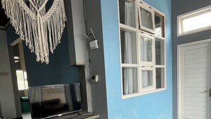 A room with a blue wall, a large window with white curtains, a macrame wall hanging, a flat-screen TV, and a colorful hexagonal rug. - Rees Cabin & Homestay (Dieng Kulon)