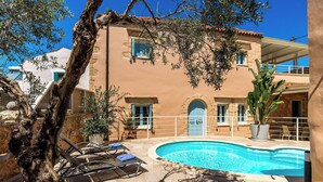 A poolside area with lounge chairs, a swimming pool, and a building with a stone facade and blue shutters. - Villa Cava – Villa with a private pool in Crete (Xirostérnion)