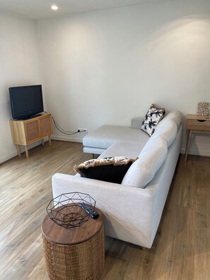 Una sala de estar moderna con un sofá de color gris claro, un televisor de pantalla plana sobre un soporte de madera, una mesita de madera pequeña y una mesita redonda de mimbre.