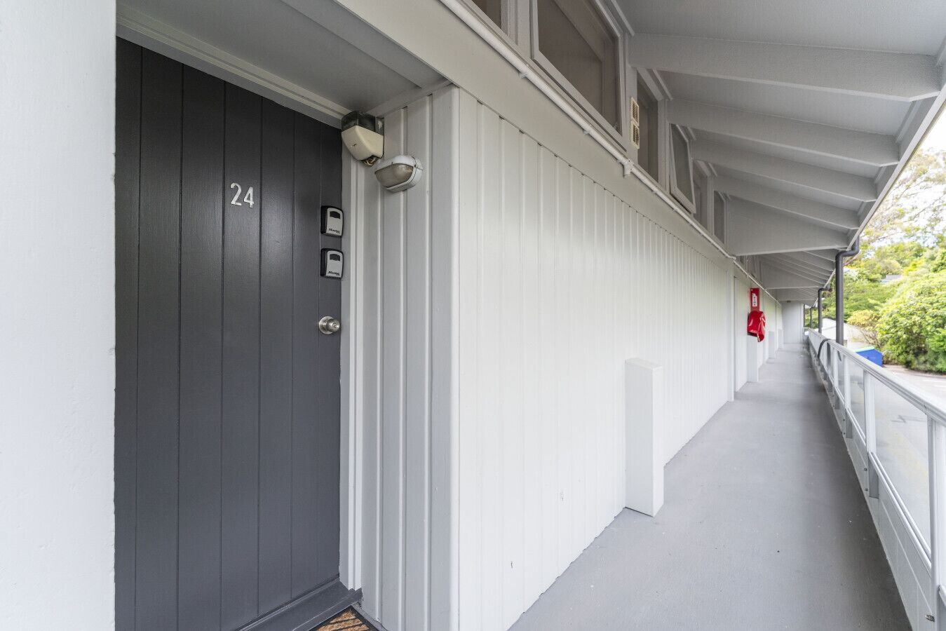 Un couloir avec une porte grise portant le numéro 24, un mur blanc et un extincteur rouge sur le côté droit.