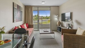A modern living room with a sofa, a coffee table, and a TV. There is a glass table with a vase and a plate of fruit. The room has a large window with a view of a road and greenery. - Lake Terrace Apartment (Taupō)