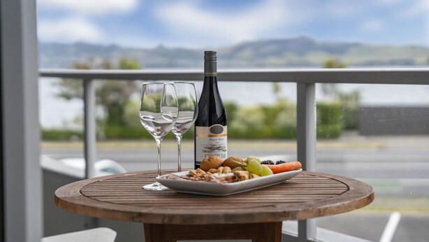 A wooden table with a bottle of wine, two glasses, and a plate of food on a balcony with a view of the mountains. - Lake Terrace Apartment (Taupō)