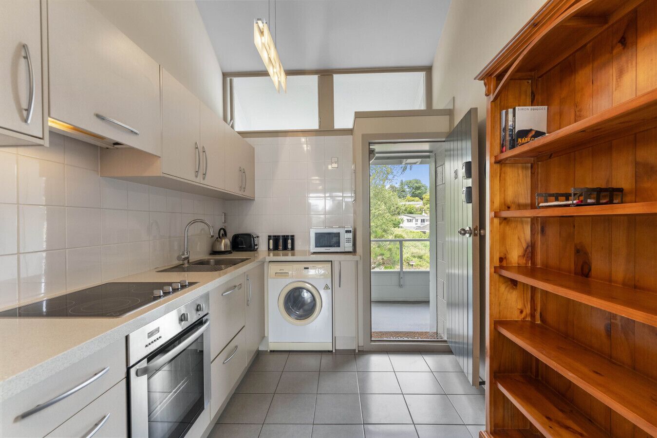 A compact kitchen with a washing machine, a microwave, and a toaster. There is a wooden shelf with books and a view of the outside.