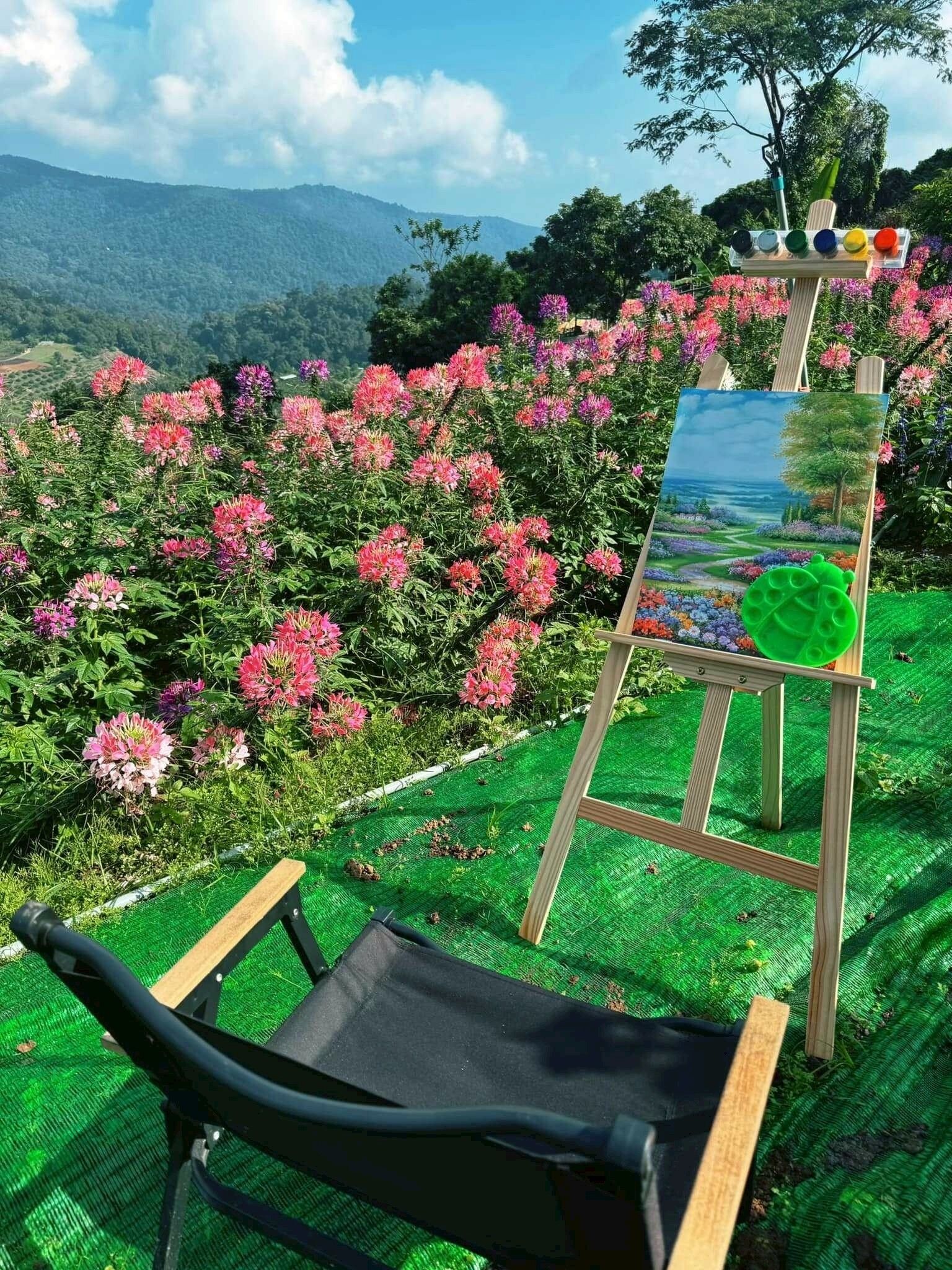 Un caballete con una pintura de un paisaje, dispuesto al aire libre en un jardín con flores rosadas y vistas a colinas distantes.