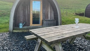 Eine gemütliche, halboffene Holzhütte mit einer Tür, einem kleinen Tisch mit einem Glas und einer blauen Wasserkanne.