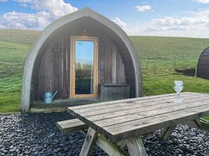 En mysig, delvis öppen träkonstruktion med en dörr, ett litet bord med ett glas och en blå vattenkanna.