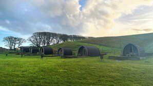 Eine Reihe von rustikalen, zylindrischen Holzhütten auf einem grasbewachsenen Hügel, mit Bäumen und bewölktem Himmel im Hintergrund.