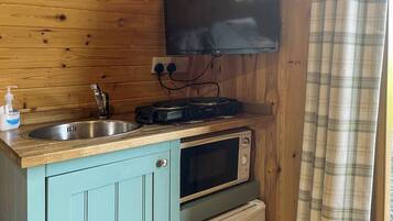 A compact kitchen with a wooden wall, a flat-screen TV mounted above the counter, a sink, a coffee machine, and a microwave.