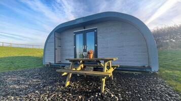 A small, semi-circular structure with a dark door and a window, a wooden picnic table in front, and a sign reading "Valley Pods."