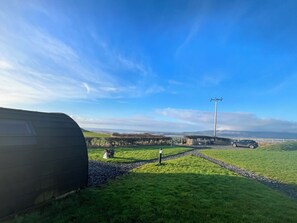 Ein grünes Feld mit einem Wohnwagen und einem geparkten Auto, einem Strommast und einer entfernten Aussicht auf Hügel und ein Gewässer.