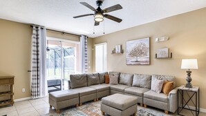 Uma sala de estar com sofá em seção, um ventilador de teto, uma mesa lateral com abajur e uma janela grande com cortinas.