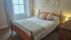 A neatly made bed with a wooden headboard, a floral painting on the wall, and a window with curtains. - 2 bedroom accommodation in Looe (Looe)