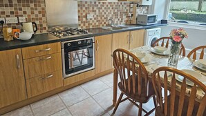 A kitchen with wooden cabinets, a gas stove, an oven, and a dining area with chairs and a table. - 2 bedroom accommodation in Looe (Looe)