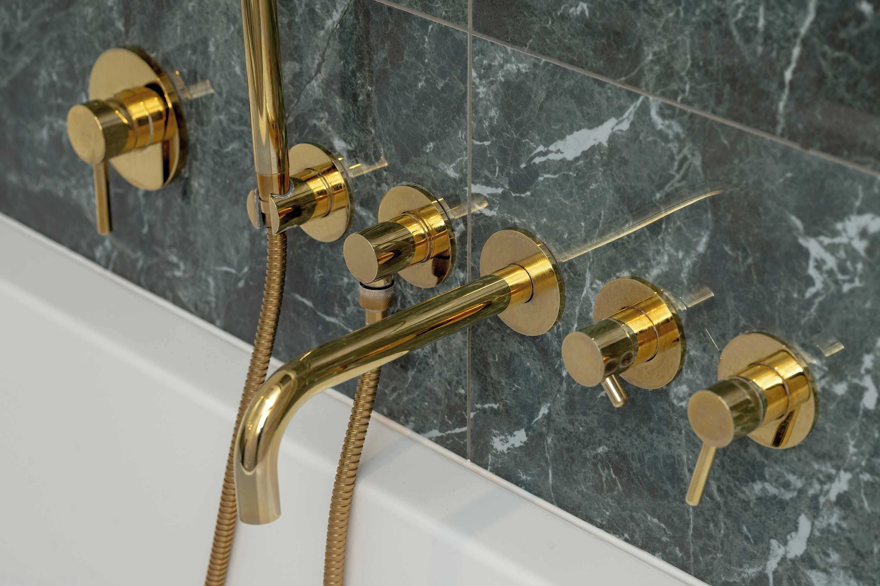 A bathroom with a gold showerhead and controls, a white bathtub, and dark marble tiles.