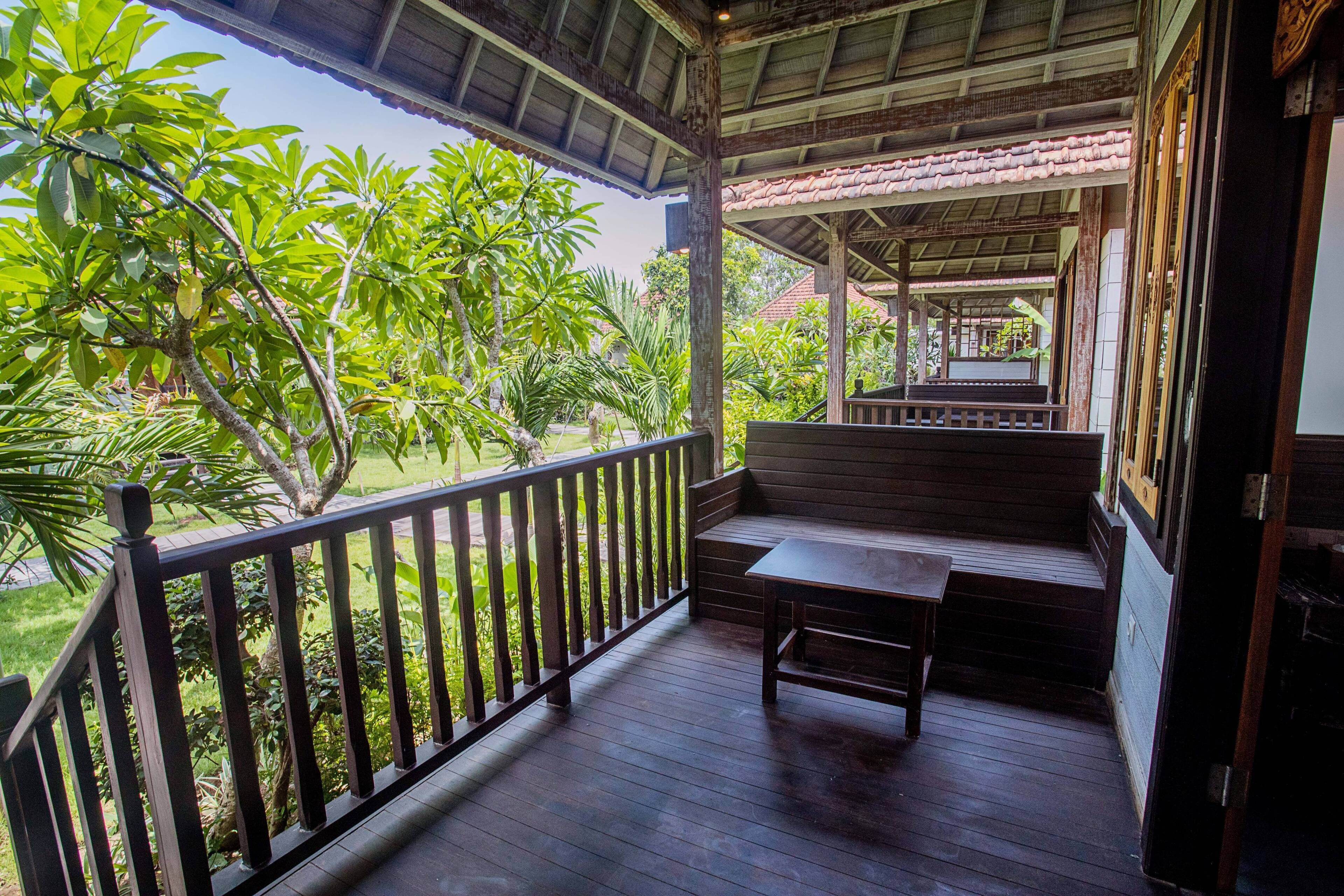 A wooden porch with a bench and railing, overlooking a garden with various plants and a building in the background.