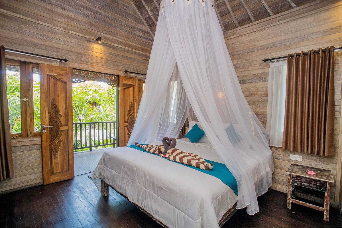 Quarto com cama de dossel, piso de madeira e varanda com vista para o verde.