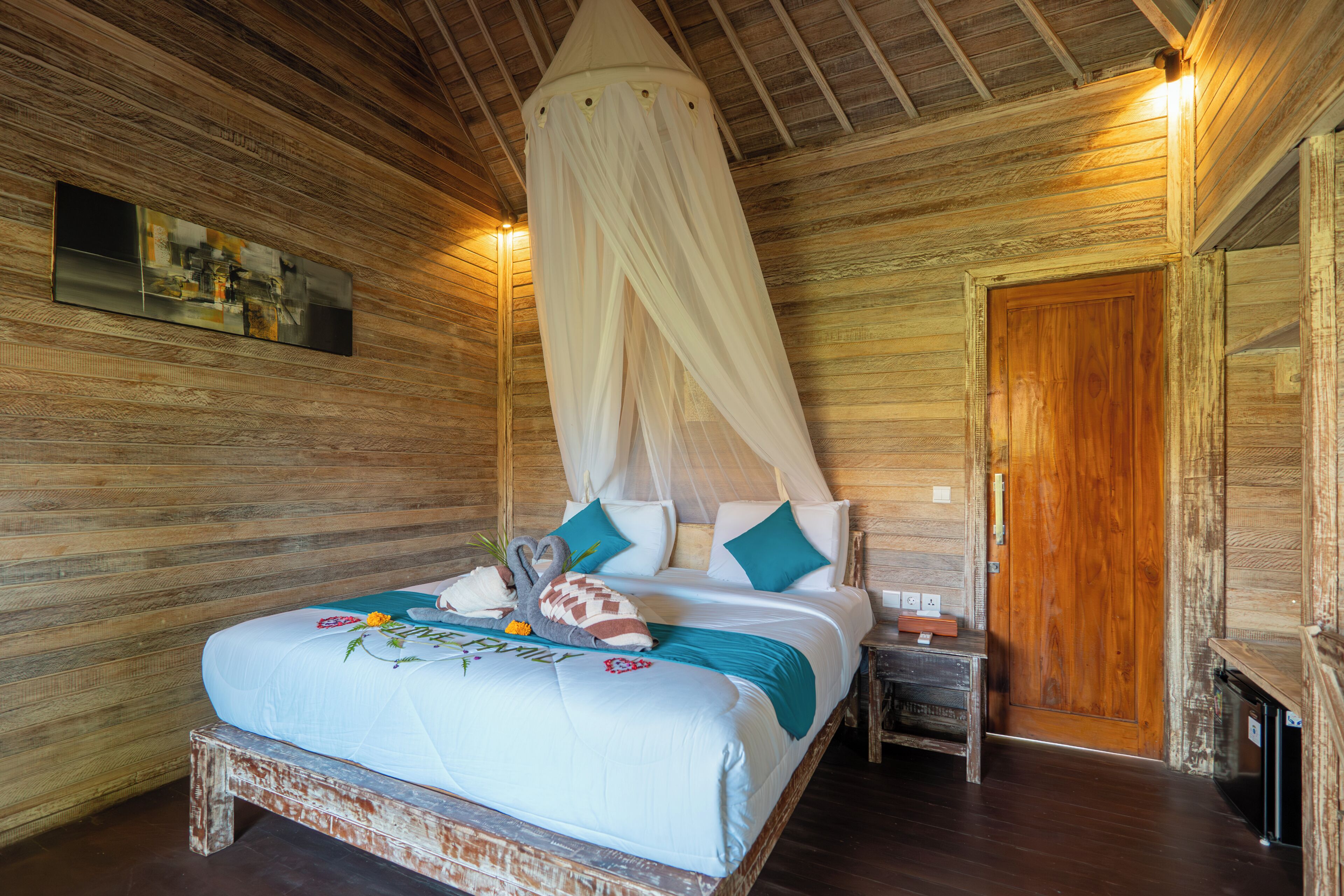 A wooden room with a bed, a bedside table, a nightstand, a wooden door, and a wall-mounted picture.