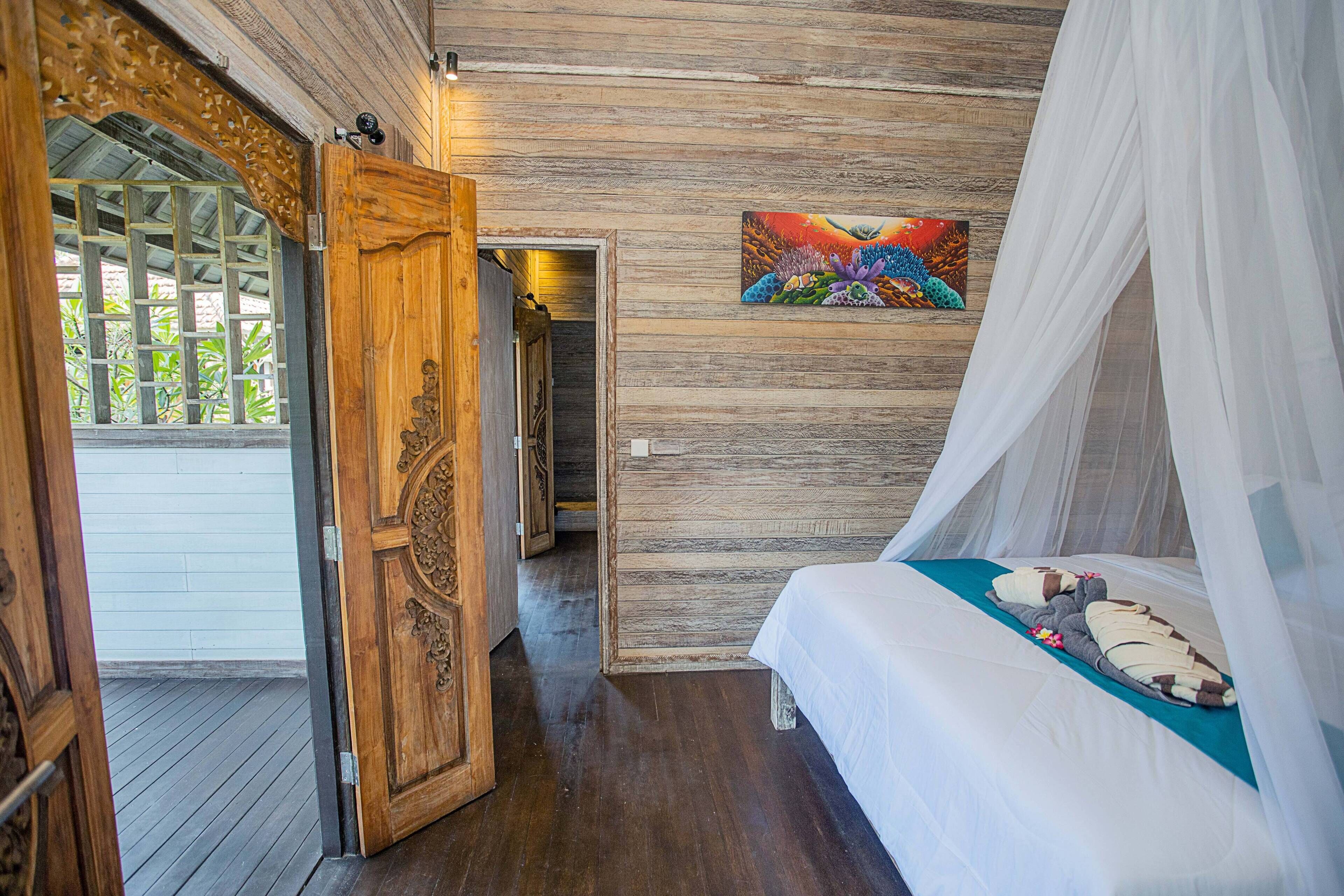 A bedroom with a wooden floor, a bed with a canopy, a painting on the wall, and a wooden door.
