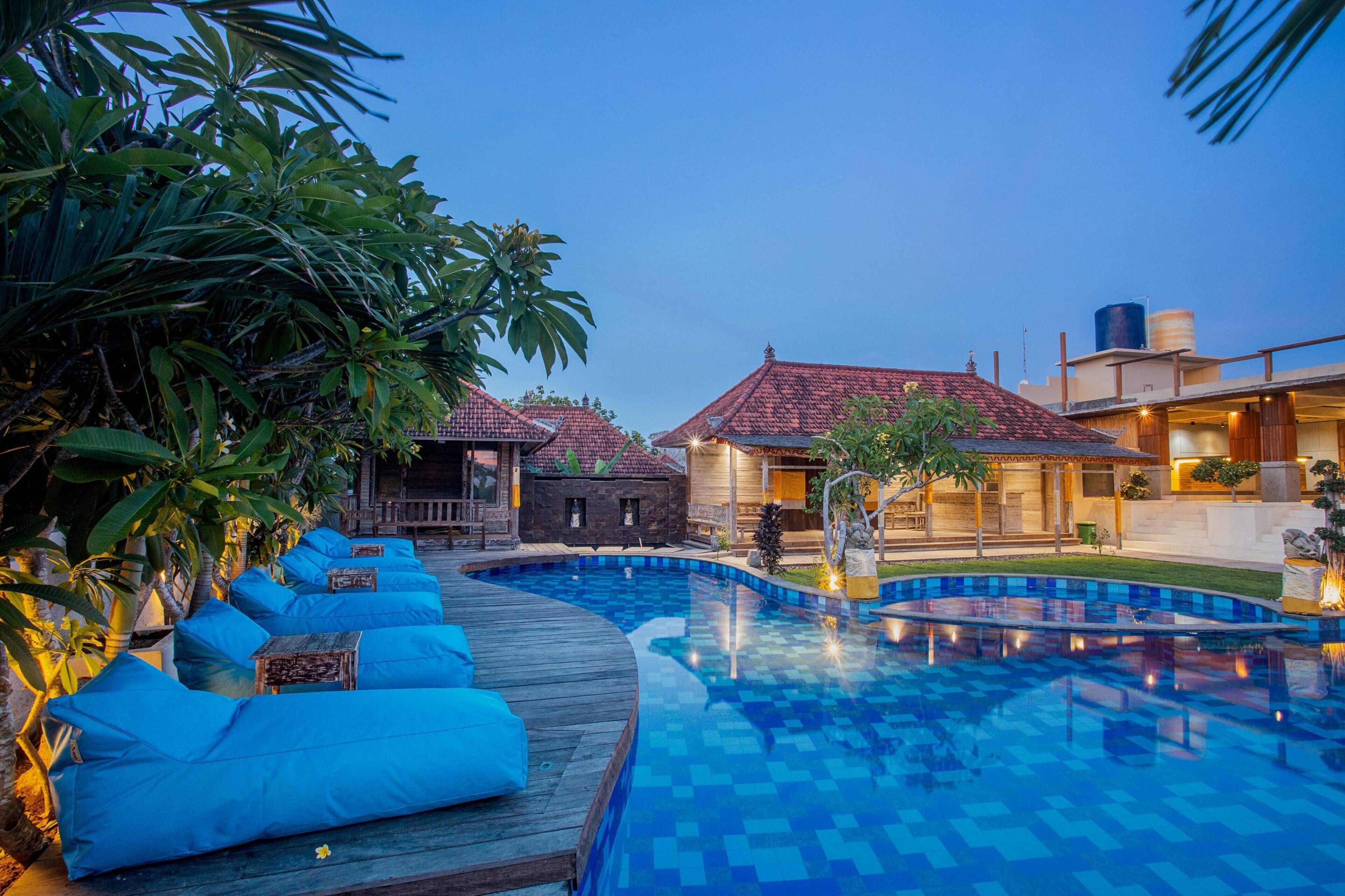 A swimming pool with a wooden deck and blue cushions, surrounded by tropical plants and buildings with red-tiled roofs.