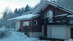 A two-story house with a red exterior, a balcony, and a garage. Snow covers the roof and ground.