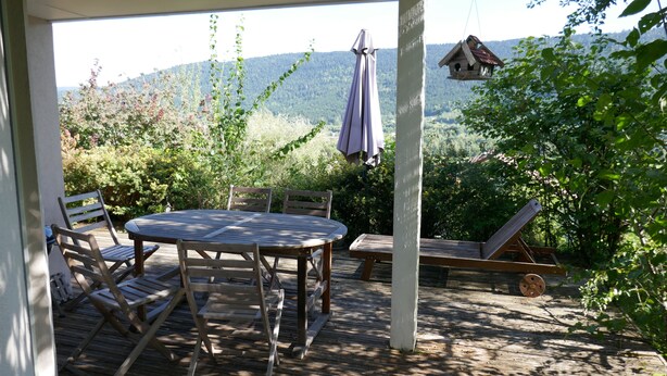 A wooden deck with a round table and chairs, a hanging birdhouse, and a view of a forested mountain range.
