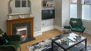 A living room with a fireplace, green armchairs, a glass coffee table, a flat-screen TV, and a chandelier. - Lakes and Shores Retreat (Maryport)
