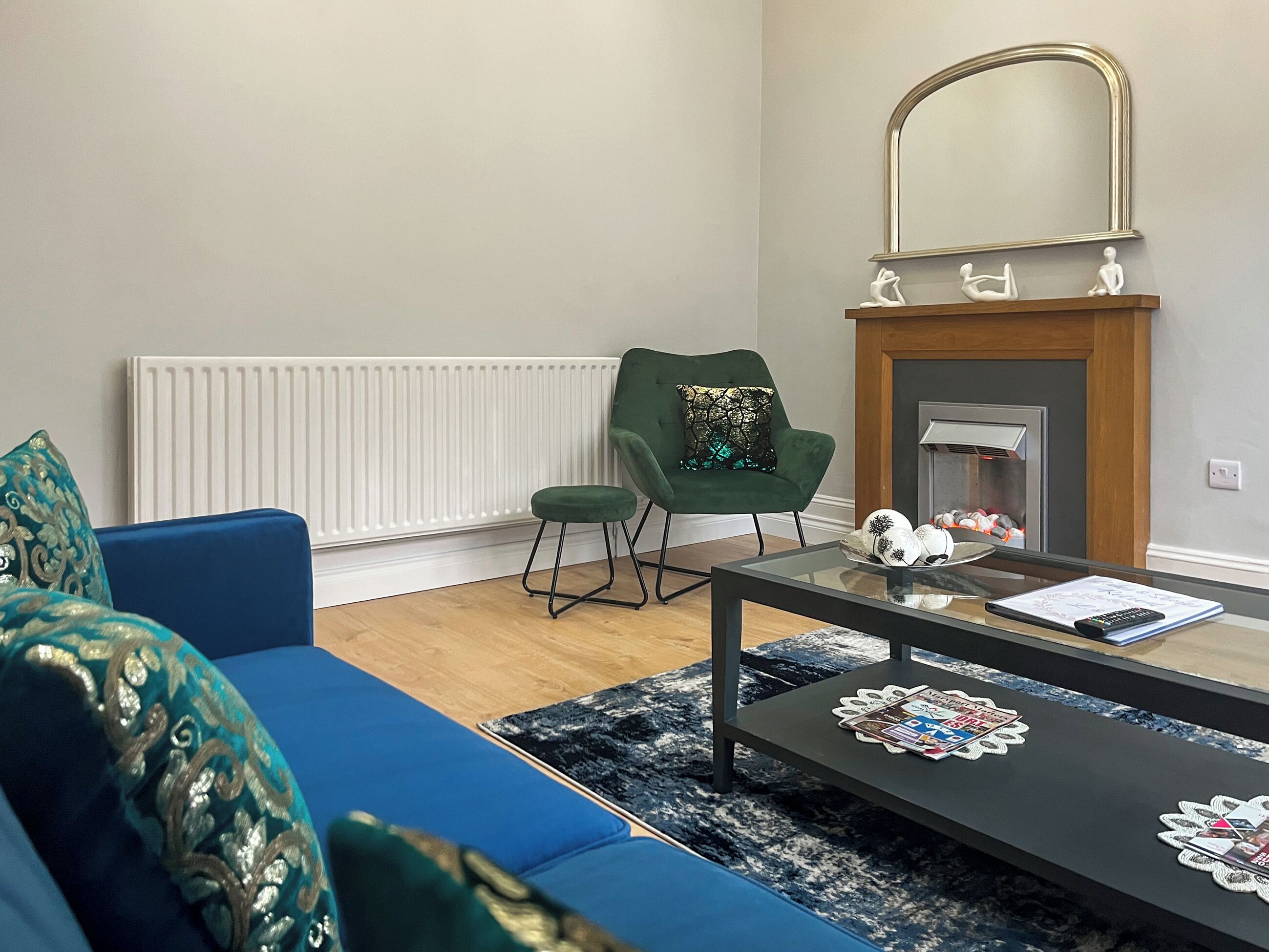 Uma sala de estar com um sofá azul, uma poltrona verde, uma mesa de centro de vidro e uma lareira decorada.