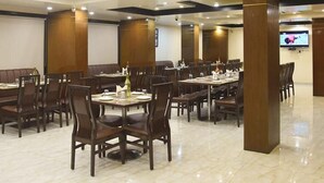 A spacious dining area with multiple tables and chairs, a large pillar, and a television screen on the wall. - Aalcajars Inn (Patna)