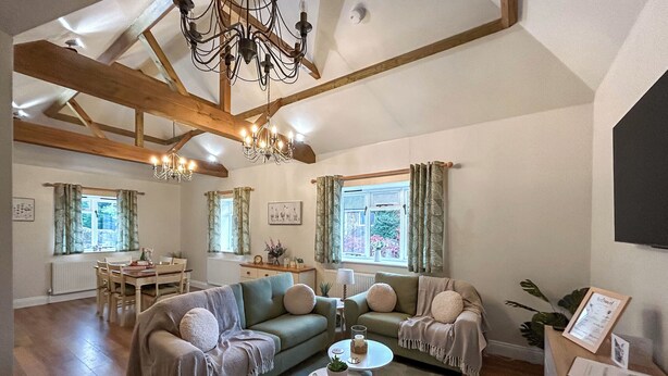 A spacious living room with a high ceiling, exposed wooden beams, a chandelier, a sofa set, a coffee table, and a dining area with a table and chairs. - Rookery Farm Holiday Cottage (Bognor Regis)