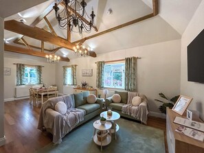 A spacious living room with a high ceiling, exposed wooden beams, a chandelier, a sofa set, a coffee table, and a dining area with a table and chairs. - Rookery Farm Holiday Cottage (Bognor Regis)
