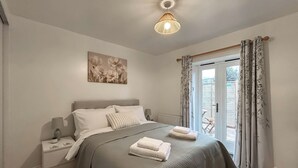 A bedroom with a bed, nightstands, a lamp, a painting, and a view through a door. - Rookery Farm Holiday Cottage (Bognor Regis)