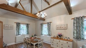 A dining area with a wooden table and chairs, a chandelier, and a view of the outdoors through windows with patterned curtains. - Rookery Farm Holiday Cottage (Bognor Regis)
