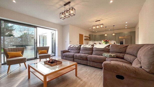 Ein modernes Wohnzimmer mit einer Couch, Sesseln, einem Couchtisch und einer Küche im Hintergrund.