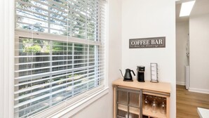 Una habitació amb finestra i persianes, una zona de bar de cafè amb cafetera i vitrina de vidre, i un cartell que diu "COFFEE BAR".