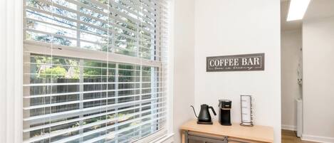 A room with a window with blinds, a coffee bar setup with a coffee maker and a glass display, and a sign that reads "COFFEE BAR".