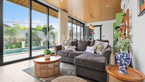 A modern living room with a grey sectional sofa, a round wooden coffee table, and a large window offering a view of a swimming pool and greenery. - Darani Villa by Kozystay - Ungasan (Ungasan)