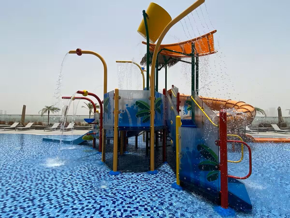 Un espace de jeux aquatiques doté de plusieurs jets d’eau et de toboggans, entouré d’une piscine dont le sol est recouvert de carreaux.