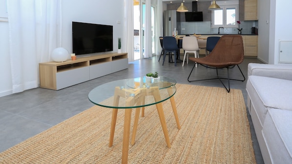 Ein modernes Wohnzimmer mit einem Glastisch, einem Flachbildfernseher, einer Couch und einem Essbereich im Hintergrund.