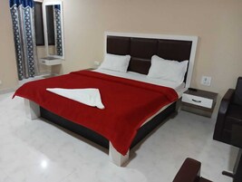Una cama bien hecha con una colcha roja y blanca, un almohadón blanco y un cabecero marrón. Hay una mesita de noche pequeña con un cajón y un espejo montado en la pared.