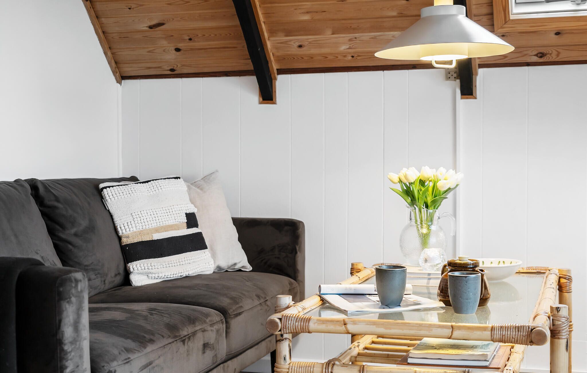 A cozy living room with a dark grey sofa, a white and black patterned throw pillow, a glass coffee table with books and decorative items, a vase with white flowers, and a hanging light fixture.