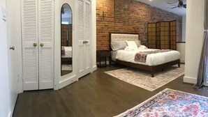 Un dormitorio con pared de ladrillo, una cama grande, piso de madera y un armario blanco con puertas batientes.