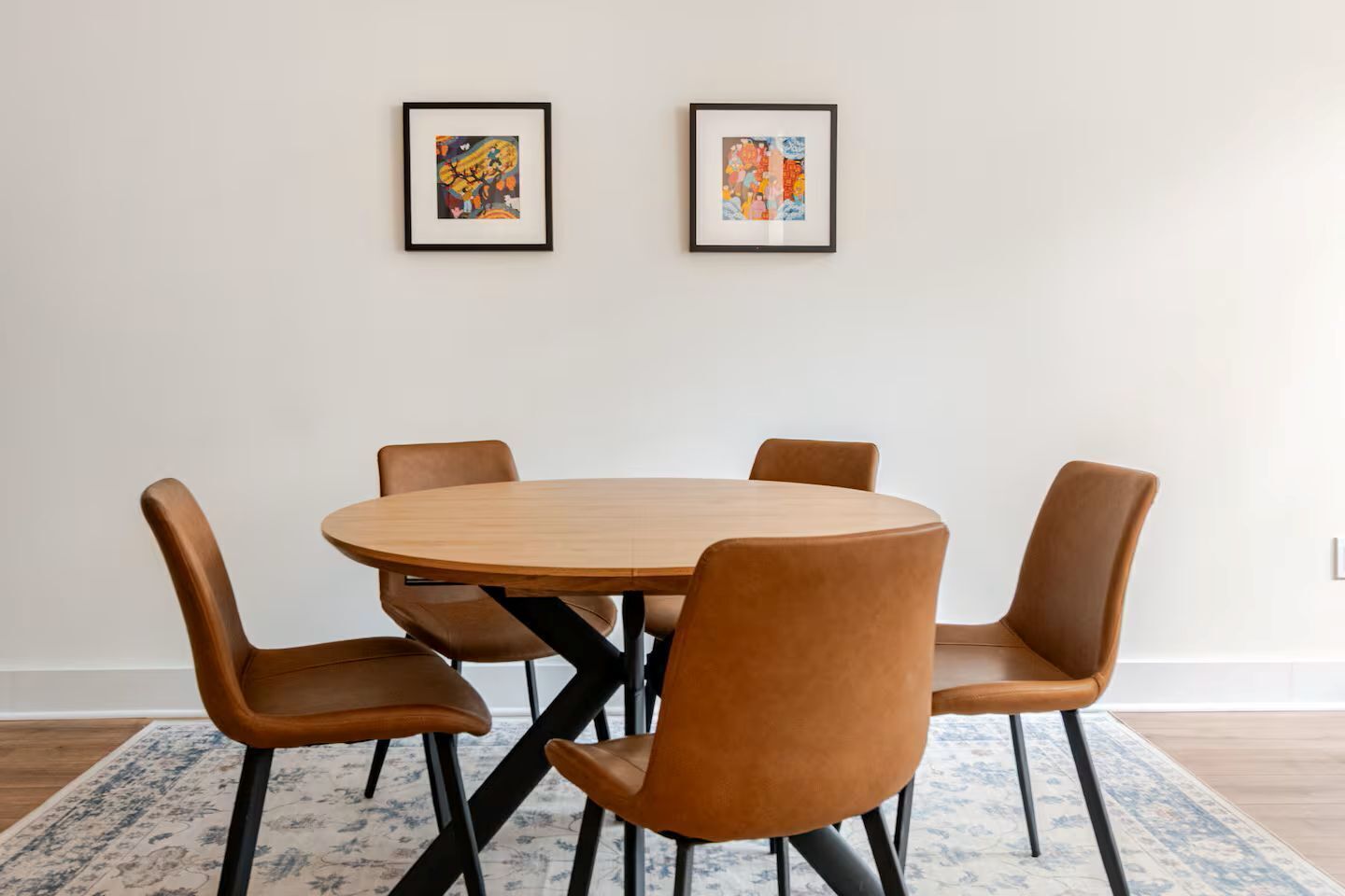 Uma sala de jantar com uma mesa de madeira redonda e quatro cadeiras marrons. Há duas obras de arte emolduradas na parede acima da mesa.