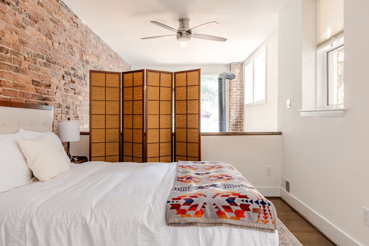 A hotel room with a brick wall, a bed with white linens, a ceiling fan, and a window with blinds.