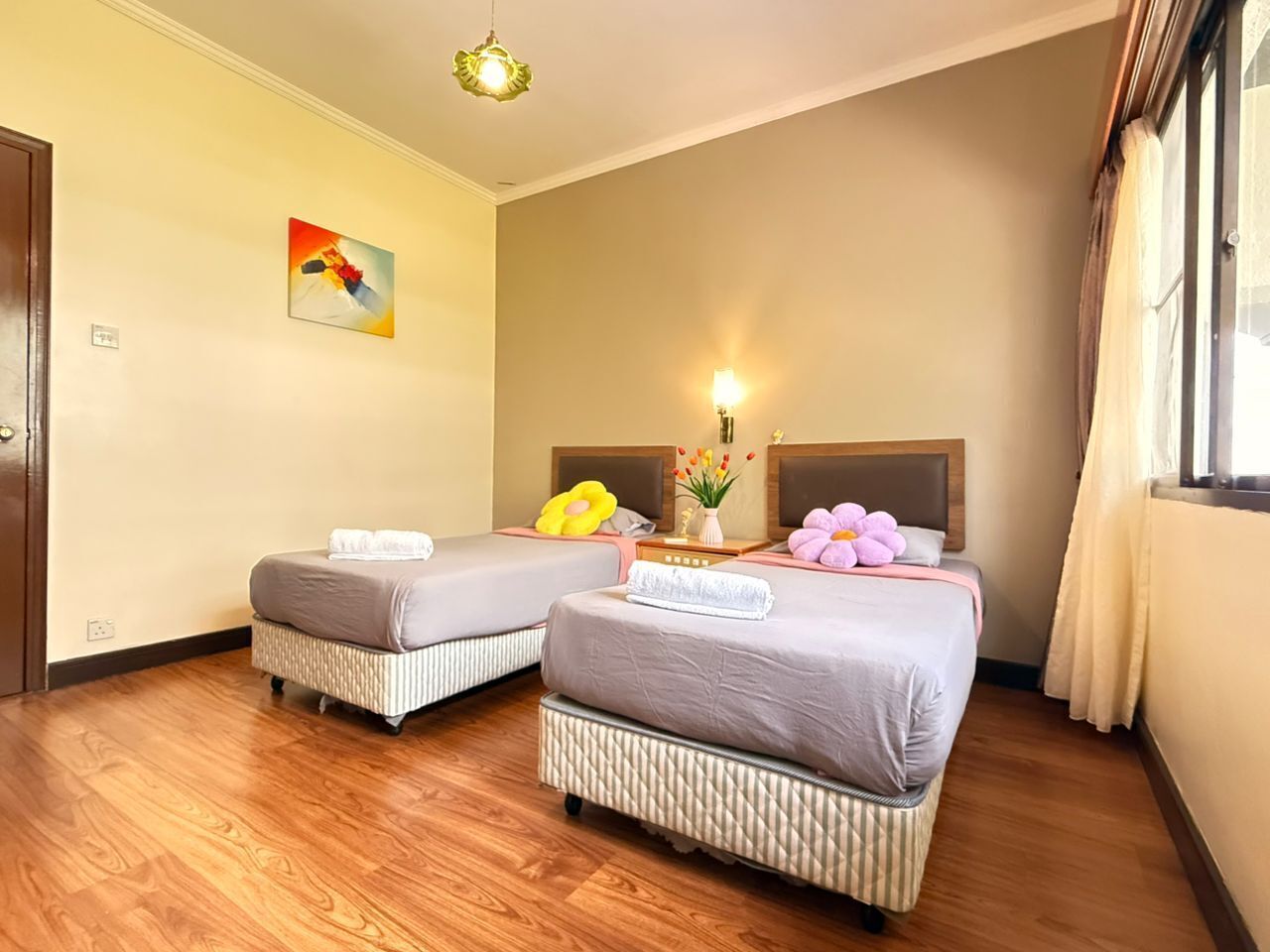 A hotel room with two beds, wooden flooring, a painting on the wall, and a window with curtains.