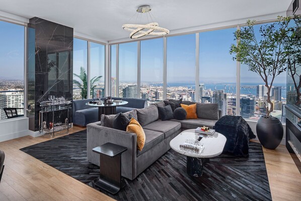 A modern living room with a large sectional sofa, a coffee table, and a view of the cityscape through large windows. - 41st Floor! 2BDR w/ Luxury Amenities & Balcony | SkyLux Penthouse by AvantStay (San Diego)