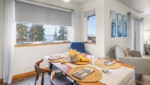 En matplats med ett bord dukat för två personer, en blå stol och utsikt över träd och en byggnad genom stora fönster.
